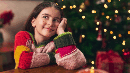 Jeune fille souriante en pull coloré assise à une table, rêveuse devant un sapin de Noël décoré et illuminé.